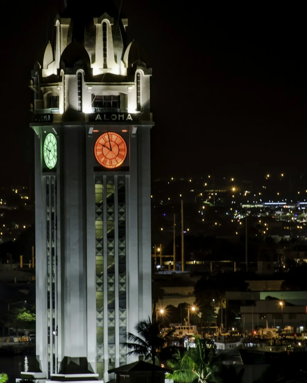 The historic Aloha Tower clock tower lit at night above Honolulu Harbor, its clock faces and "ALOHA" lettering illuminated against the dark sky